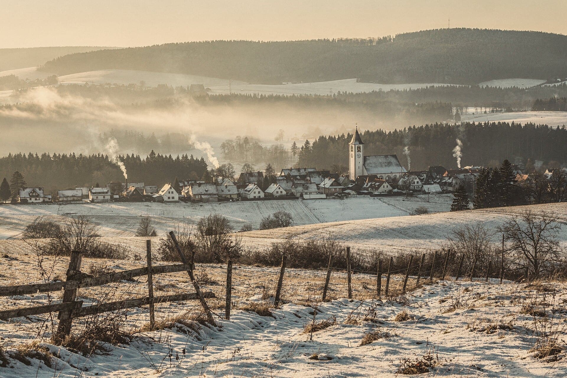 Winter in the Erzgebirge: snowy fields and spruce forest roll toward a village with a white church tower, mist in the valley.