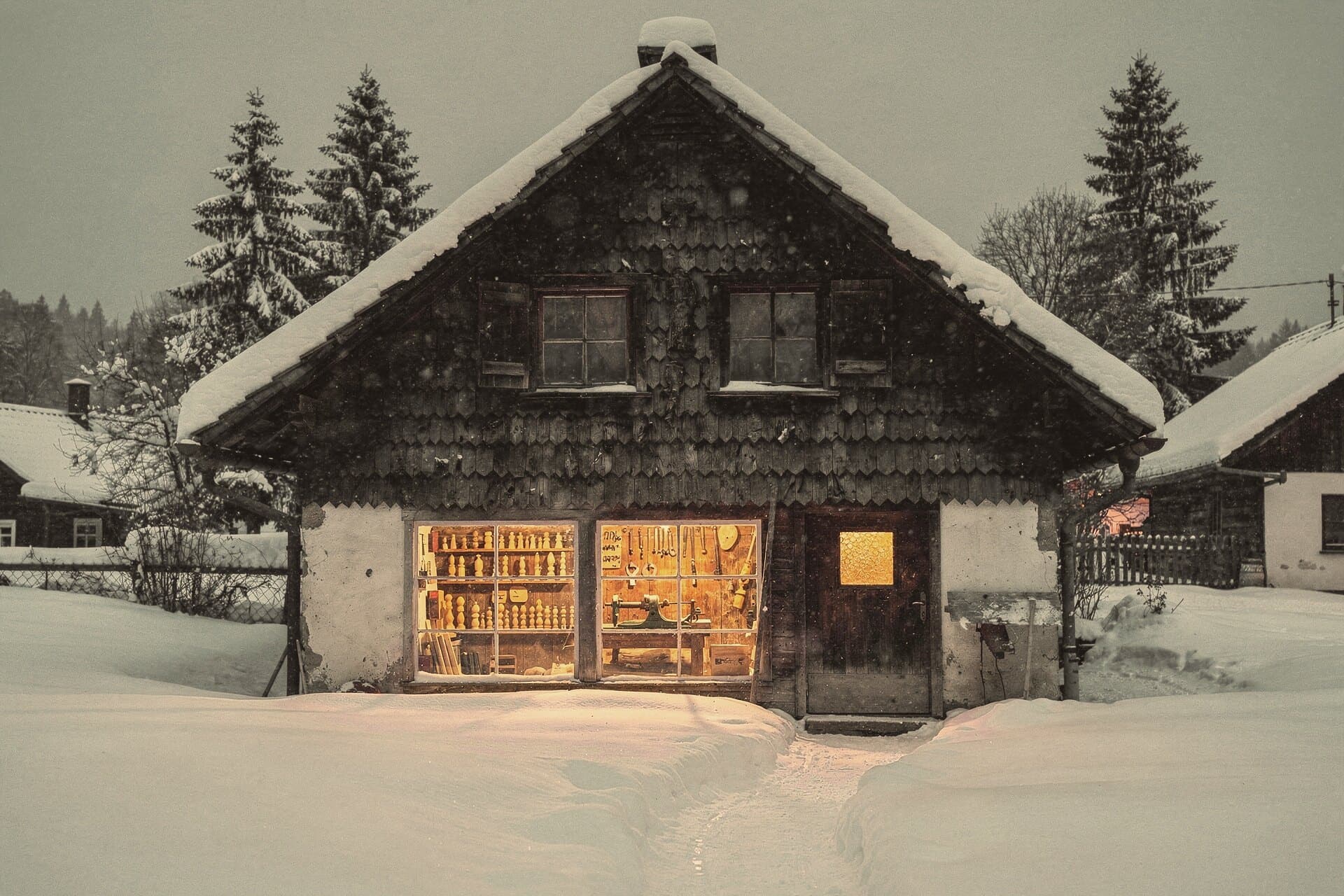 A shingled house in deep snow at dusk, its workshop windows glowing, shelves of wooden figures and a lathe inside.